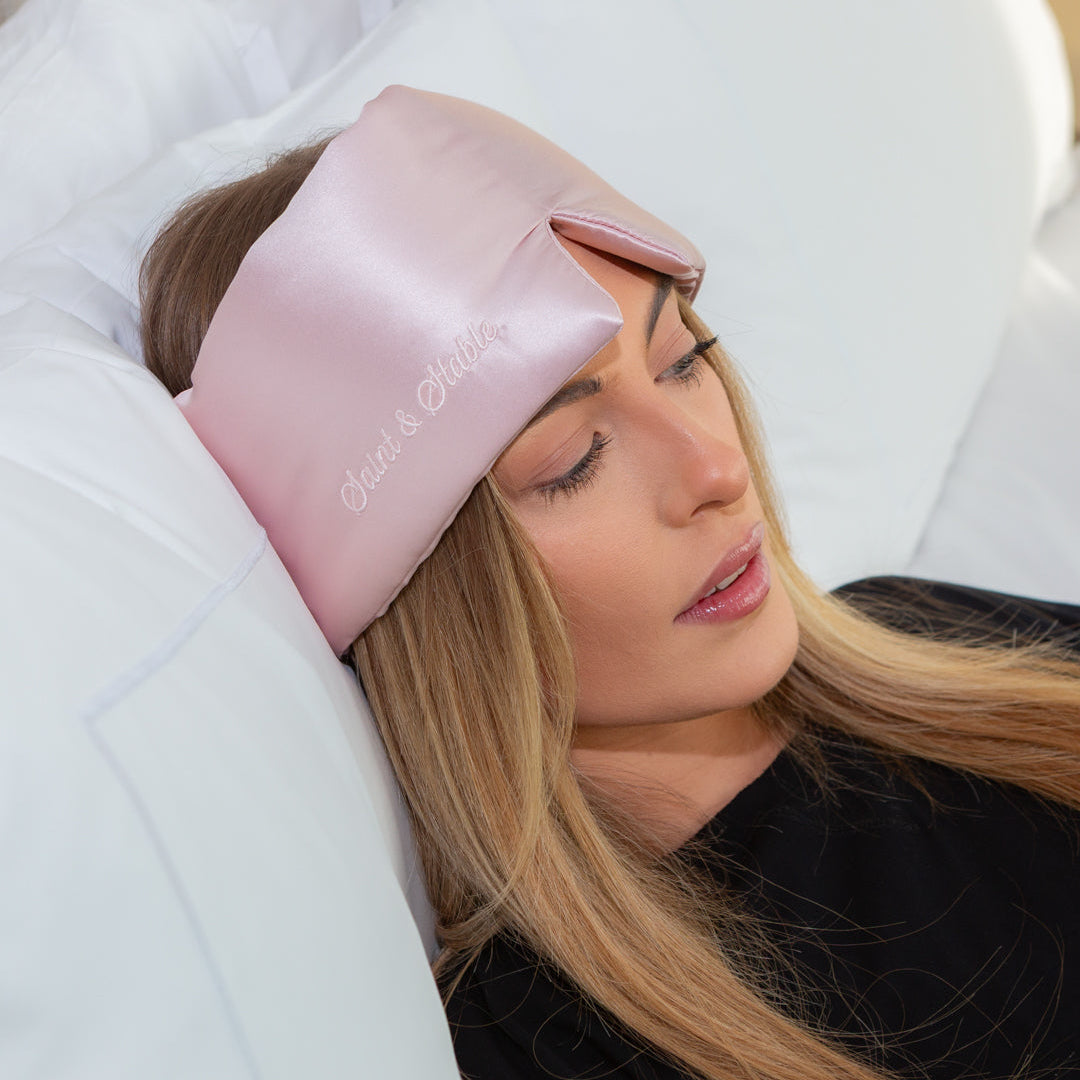 Woman wearing a pink satin eye mask with 'Sleepy' branding on a white pillow.